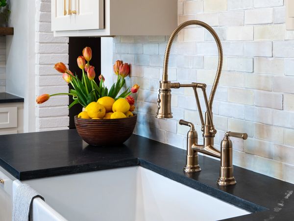 A brass kitchen faucet, with a natural white zellige backsplash, a brass faucet, and black counters