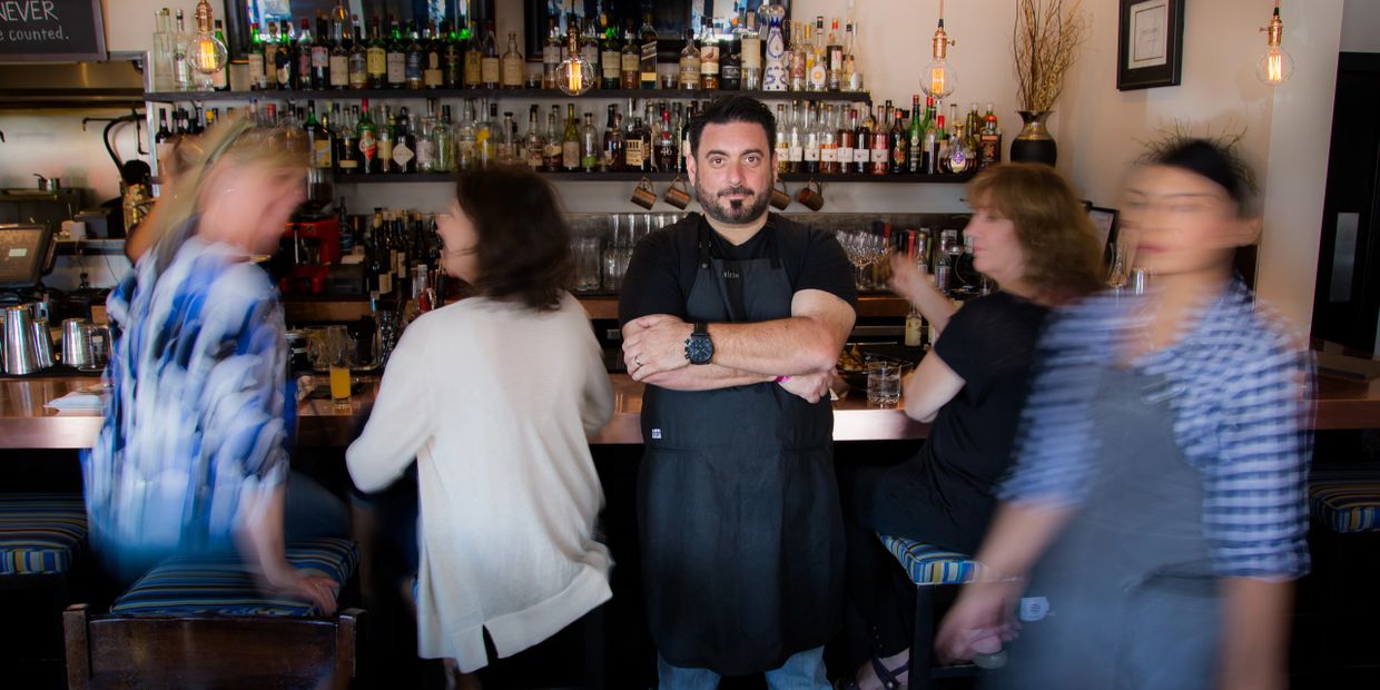 Bartender stands confidently behind a busy bar with blurred patrons around.