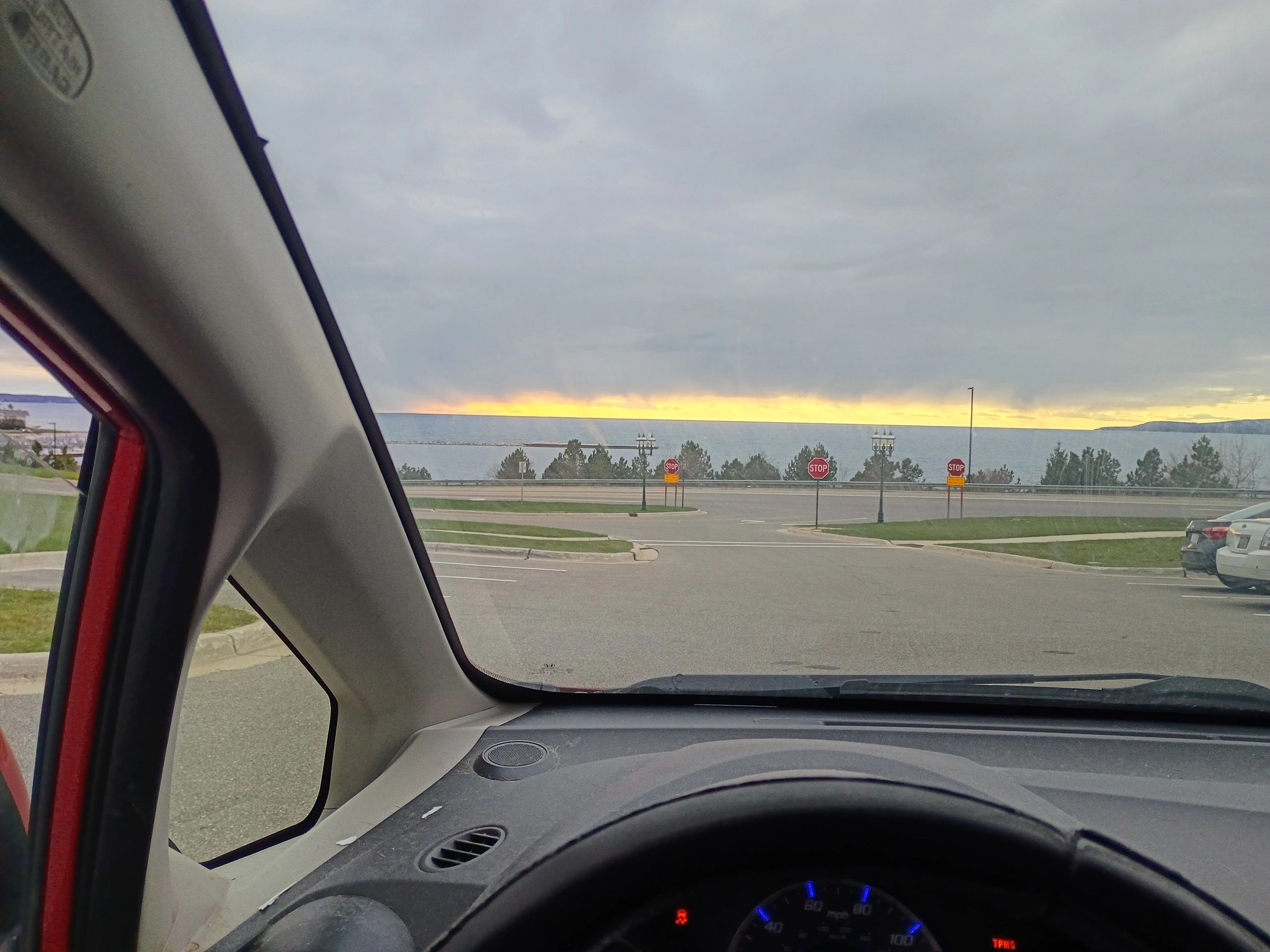 View of a sunset over water from inside a car with a stop sign ahead.