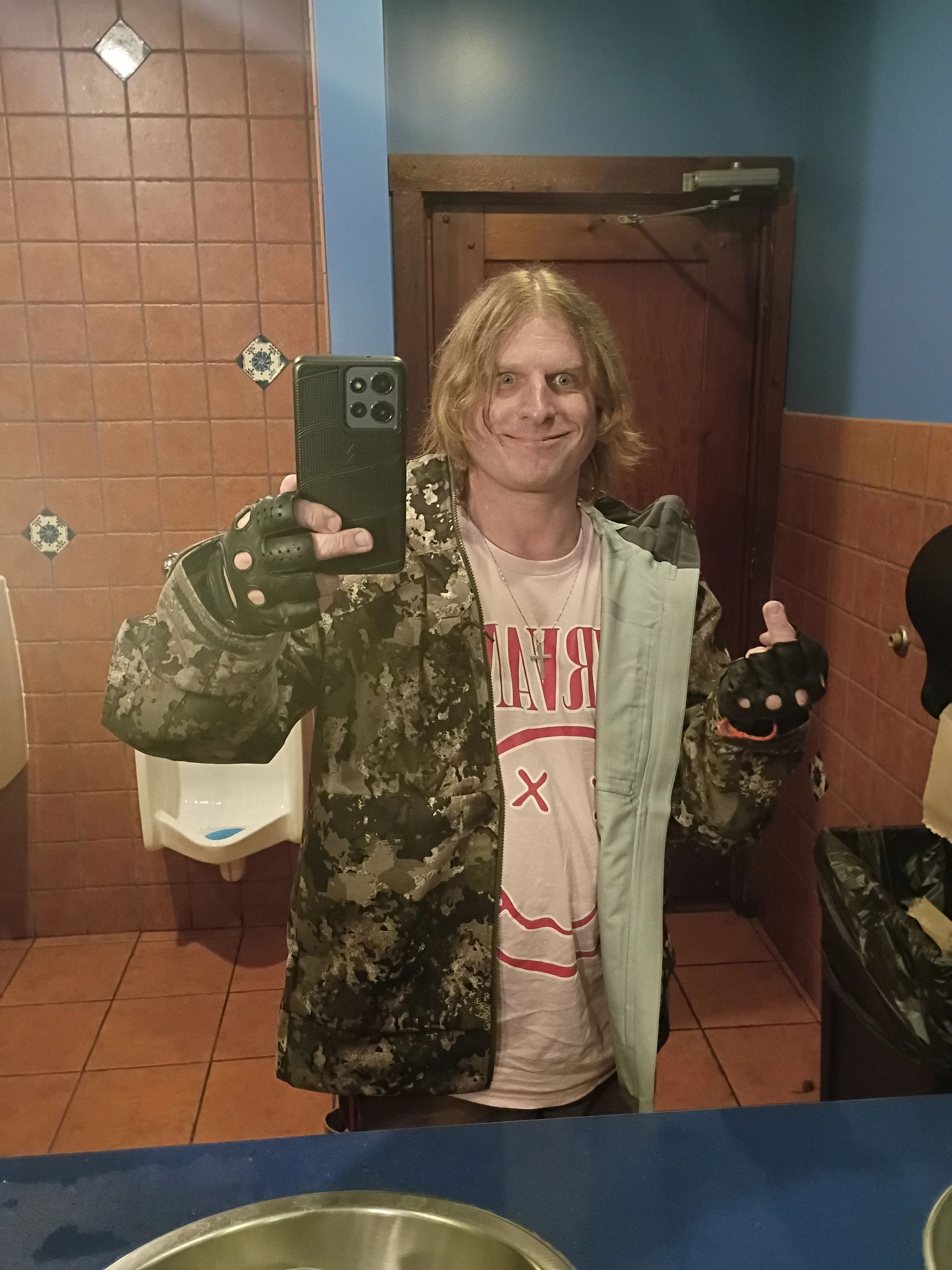 Person in camouflage jacket and gloves taking a mirror selfie in a tiled bathroom.