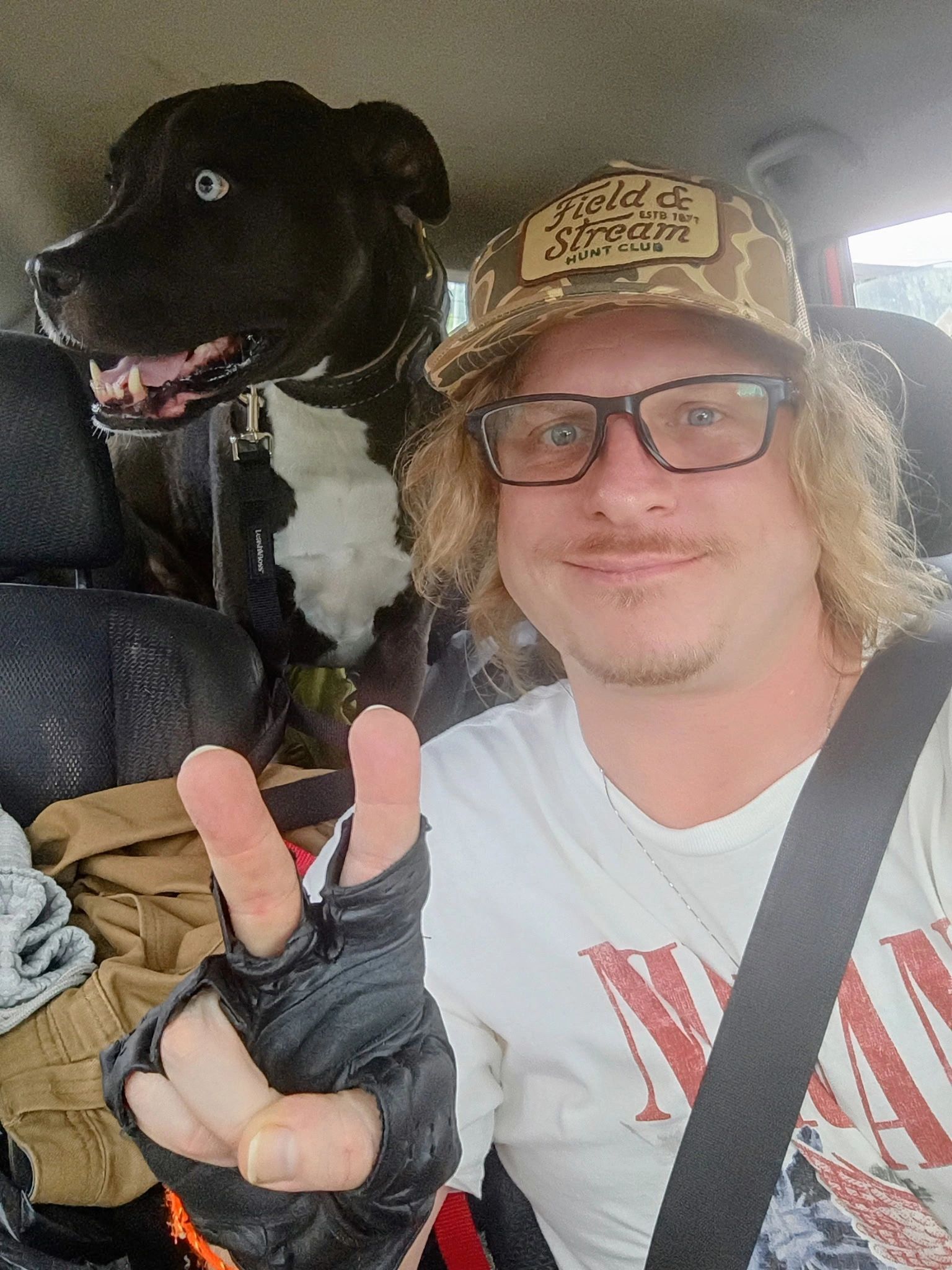 Smiling man with glasses and dog in car showing peace sign.
