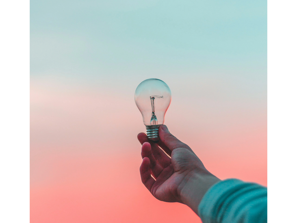 Hand holding a light bulb against a pastel sunset sky.