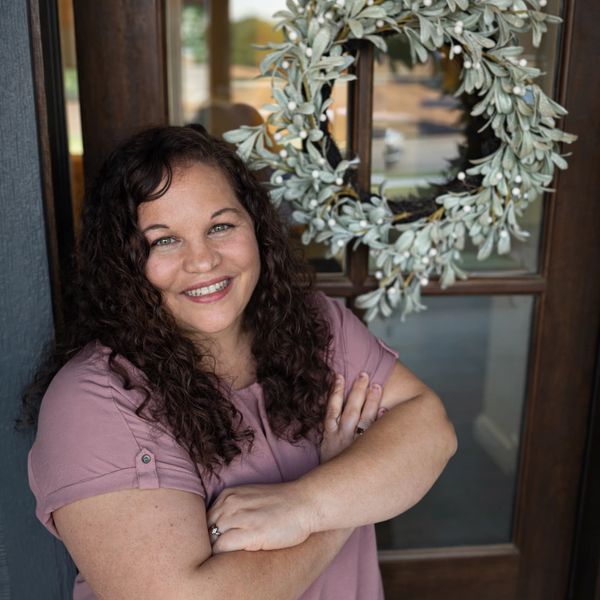 A smiling woman with curly hair stands confidently by a door with a decorative wreath.