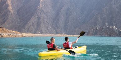 Two men kayaking on clear blue water near rocky cliffs.
