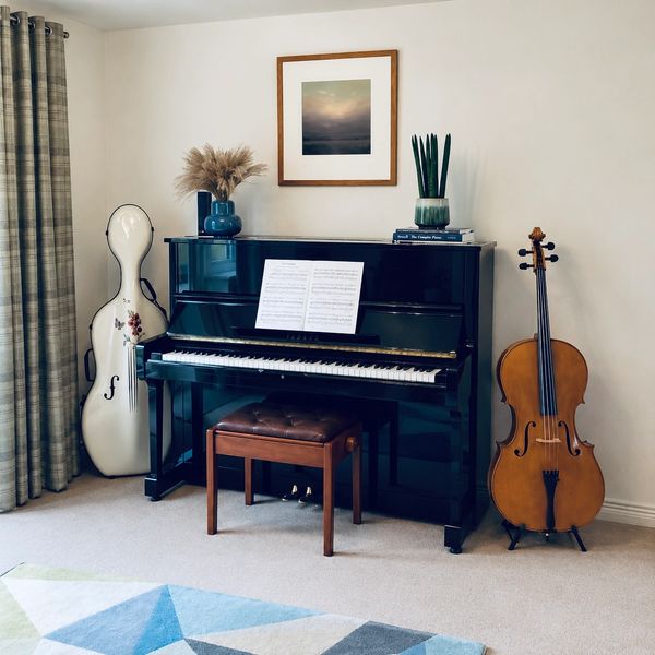 A calm, welcoming cello-teaching space featuring a black upright piano, a cello and case, framed lan