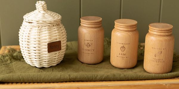 Wicker and wooden jars labeled honey, jam, and peanut butter on a cloth.