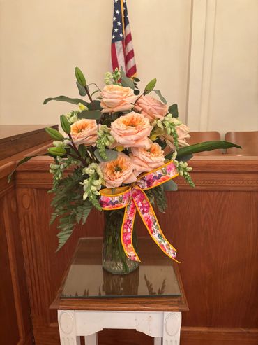 A bouquet of peach roses with greenery and a colorful ribbon in a glass vase.