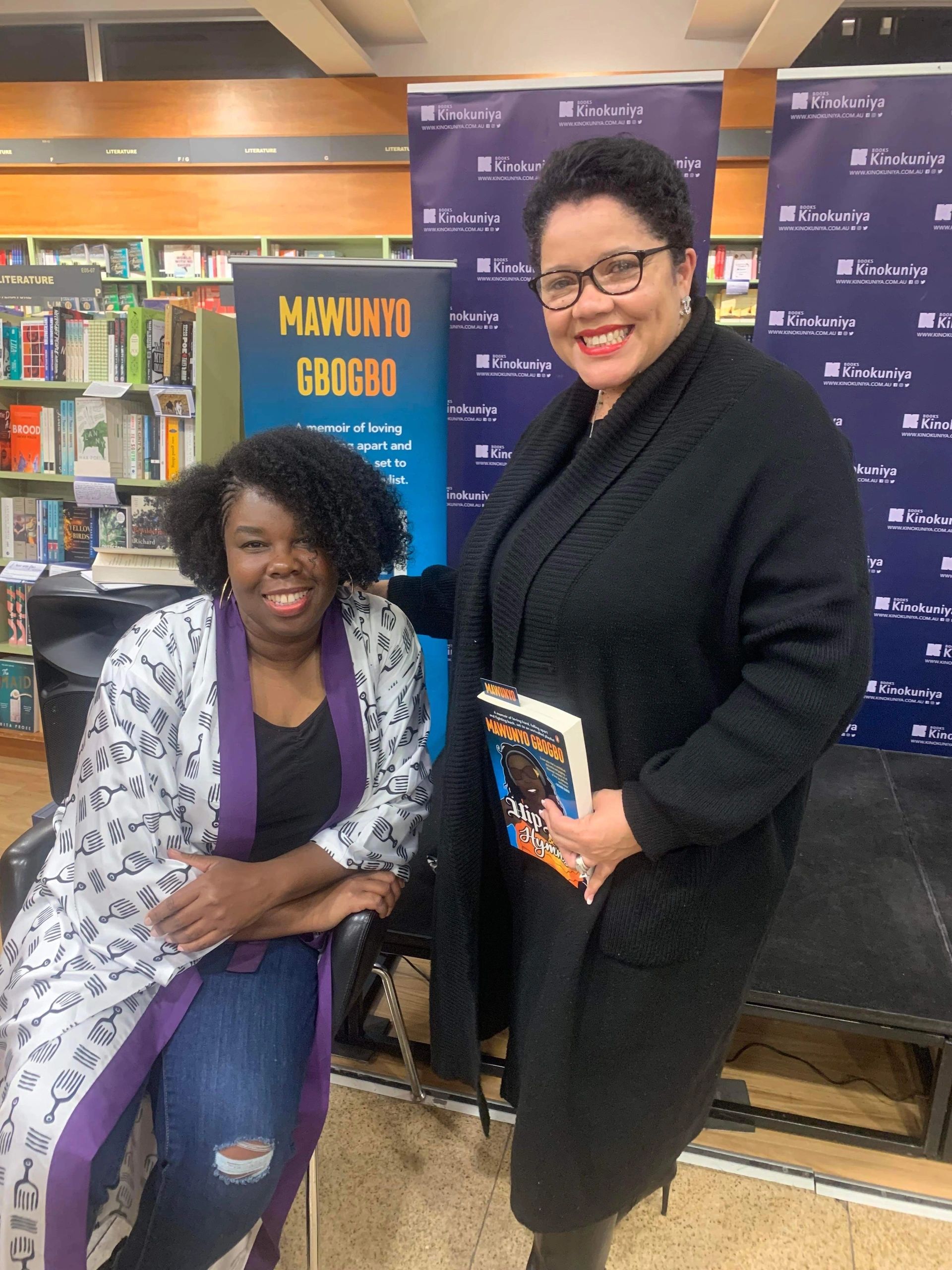 Two Black women smiling. One sitting, one standing.