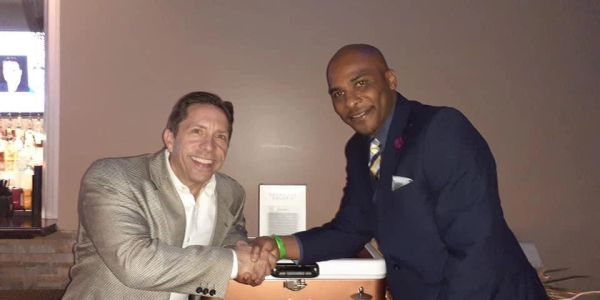 Two men in suits shaking hands and smiling indoors.