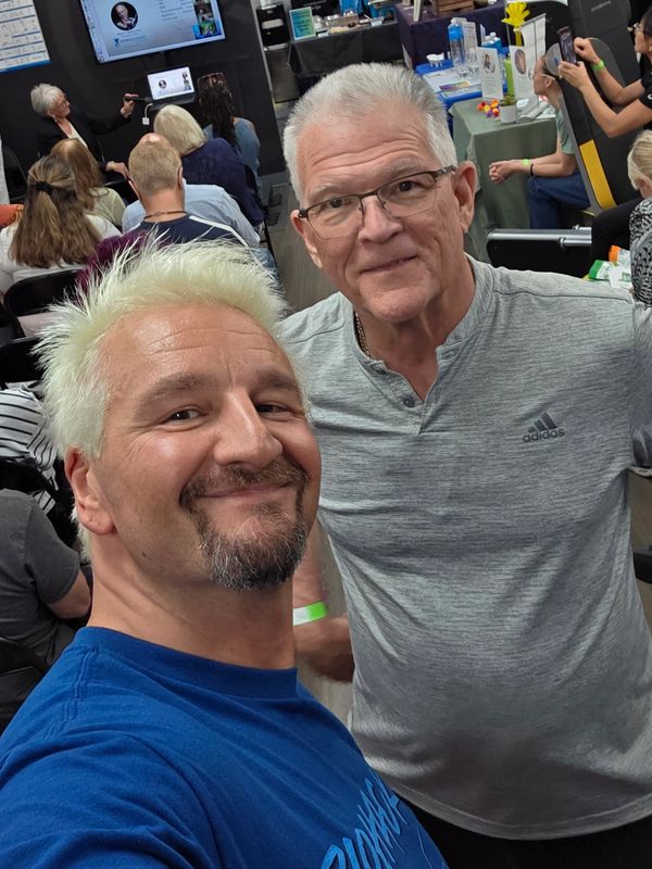 Two men smiling for a selfie in a busy indoor event with people seated and booths.
