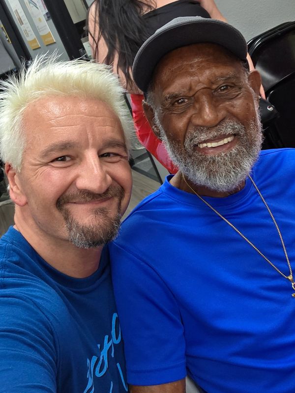 Two men smiling, one with platinum blonde hair, the other with a gray beard and blue shirt.