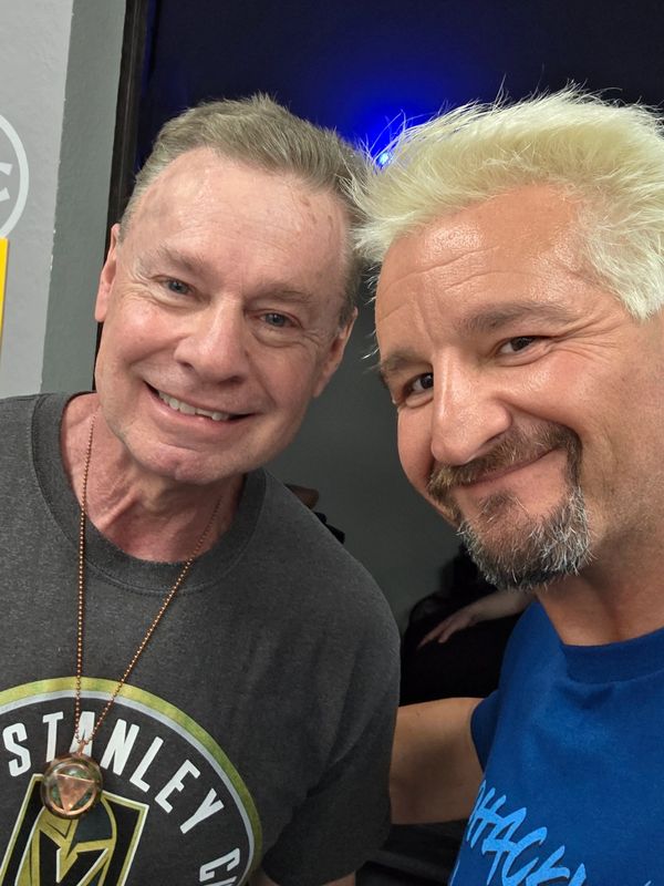Two men smiling for a selfie indoors, one with gray hair and the other with bleached blonde hair.