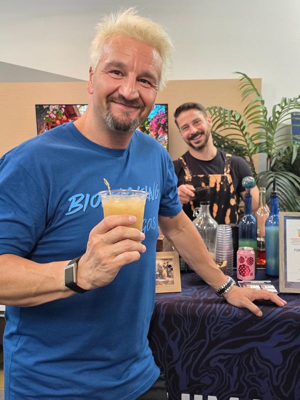 Two men smiling, one holding a drink, at a table with bottles and a can.