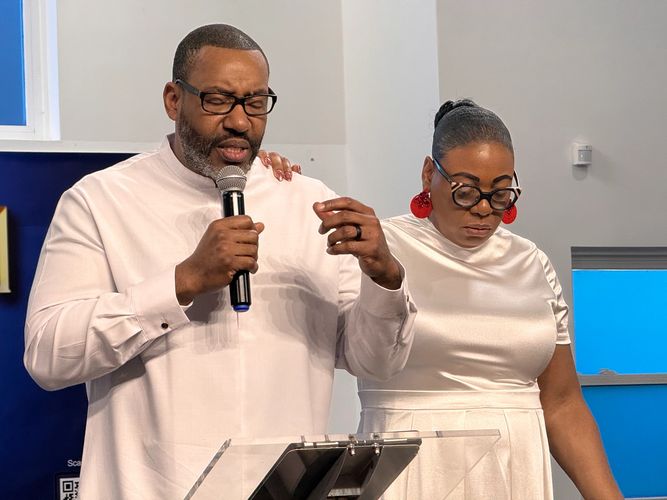 A man speaks into a microphone while a woman stands beside him with her hand on his shoulder.