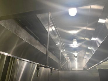 Industrial stainless steel kitchen ventilation system with hanging pipes and lights.