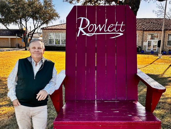 A man stands next to a giant red wooden chair labeled 'Rowlett' on a sunny day.