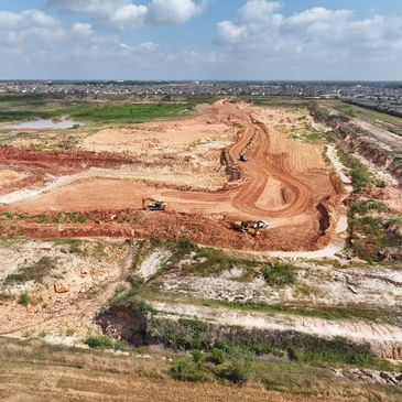 Aerial view of a large construction site with earthmoving equipment and suburban houses in the background.
