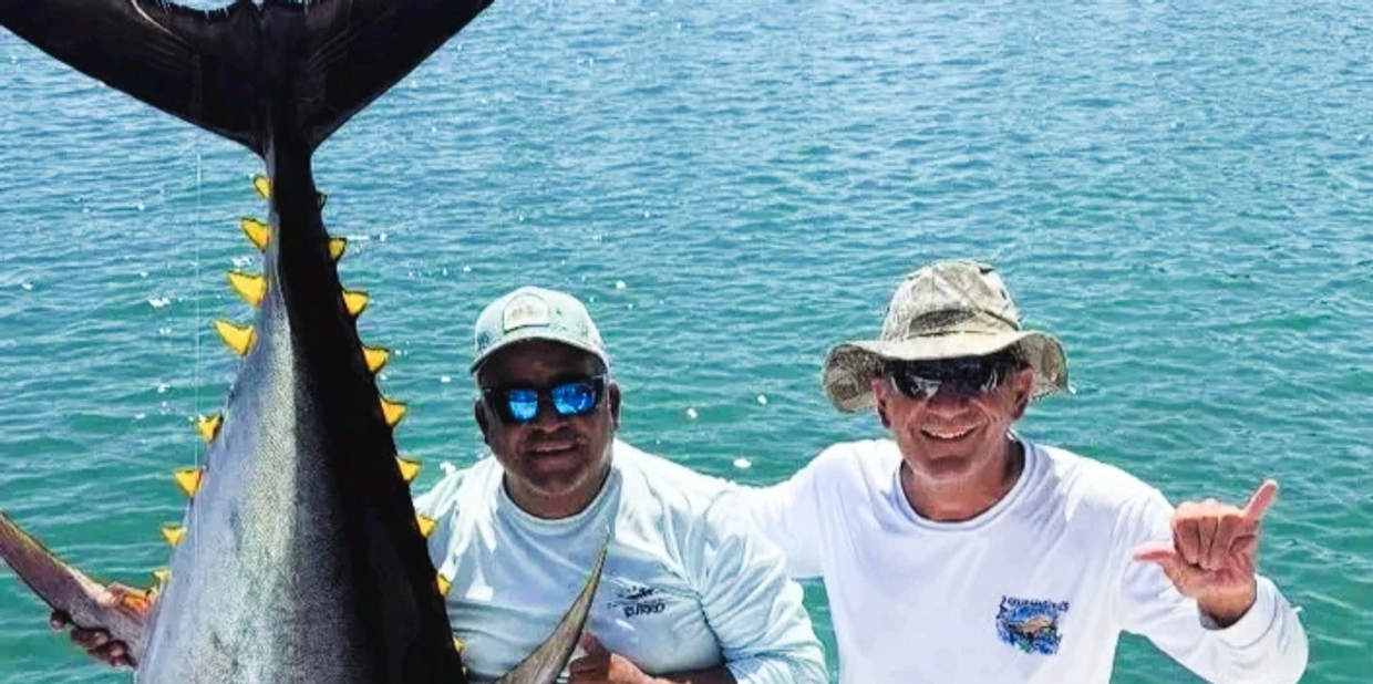 Two fishermen proudly display a huge tuna by the ocean.