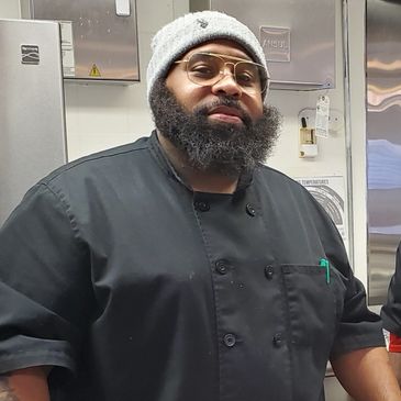 Man with beard, glasses, and hat, smiling wearing a black chef coat with pen inside of pocket