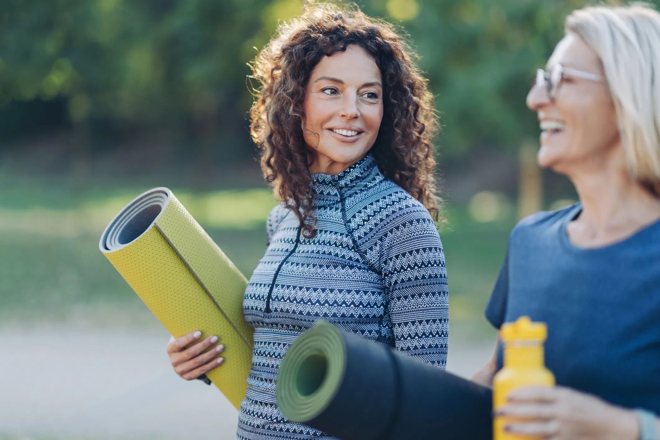 Two women smiling outdoors holding yoga mats.