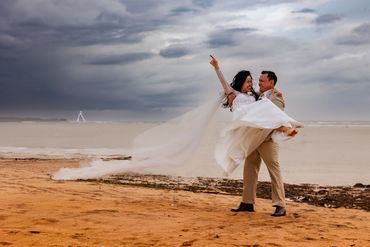 Bride in white dress lifted by groom on a beach under a cloudy sky.
