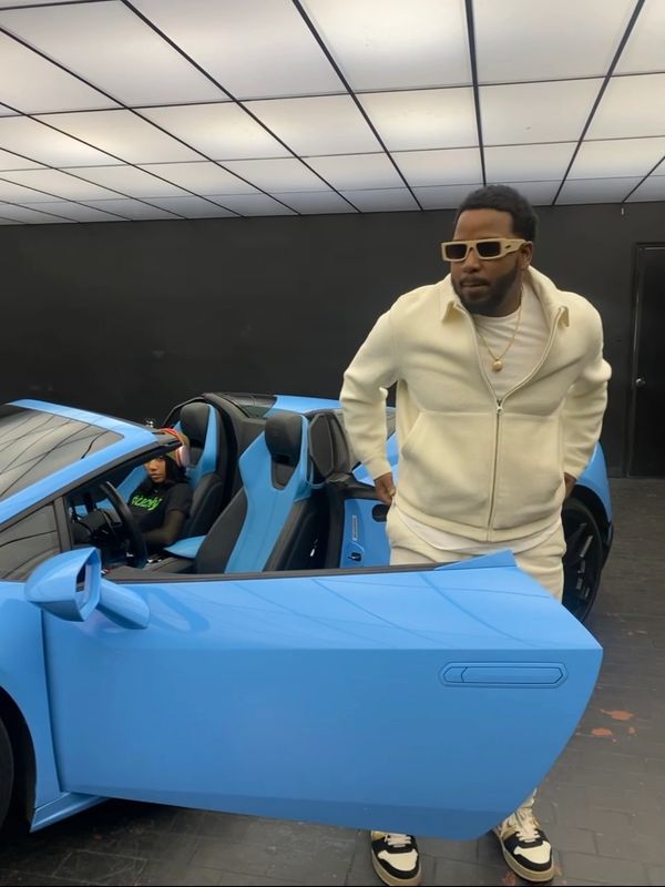Man in white outfit standing by blue sports car with woman inside.