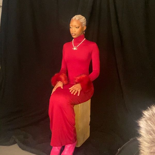 Person in a red dress with fur cuffs sitting on a wooden box against a black background.