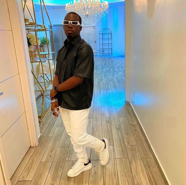A stylish man poses indoors in a black shirt, white pants, and white sneakers under a chandelier.