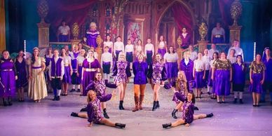A vibrant stage performance with dancers and a large chorus in purple costumes.