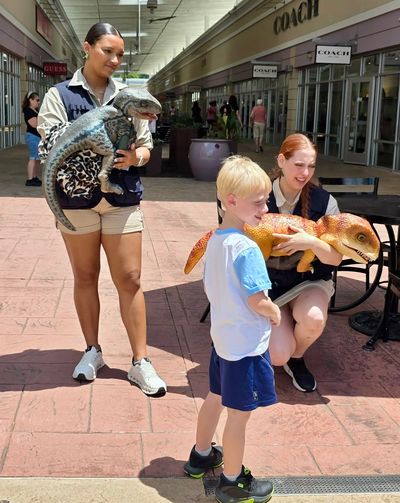 Two women hold realistic dinosaur puppets while a young boy watches in a shopping area.