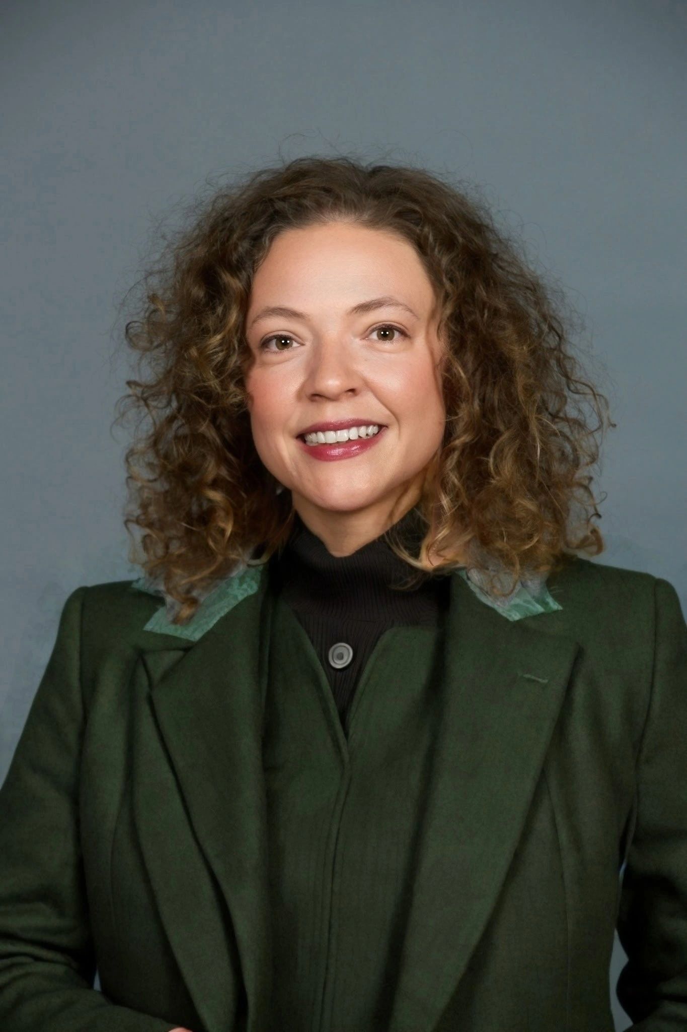 Smiling woman with curly hair wearing a green coat against a gray backdrop.