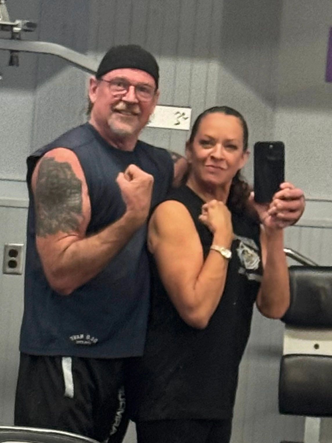 Two people flexing muscles in a gym mirror selfie.