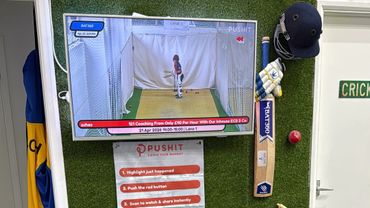 Cricket training setup with TV showing bowling practice and cricket gear on wall.