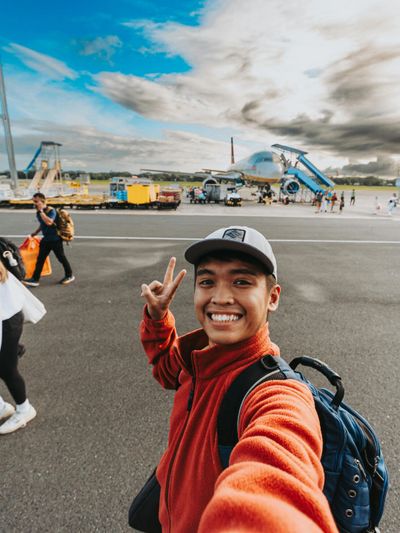 Young man takes a cheerful selfie at the airport runway with an airplane in the background.