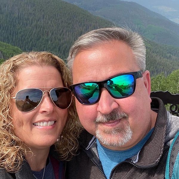 Couple wearing sunglasses smiling outdoors with forested mountains in the background.