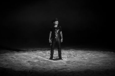A man in leather stands alone in a spotlight on a dark stage.