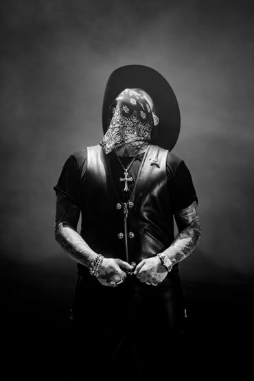 Person in a hat and bandana with tattoos and jewelry in dramatic black and white.