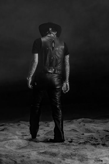 Man in leather outfit and hat standing on sandy ground, facing away.