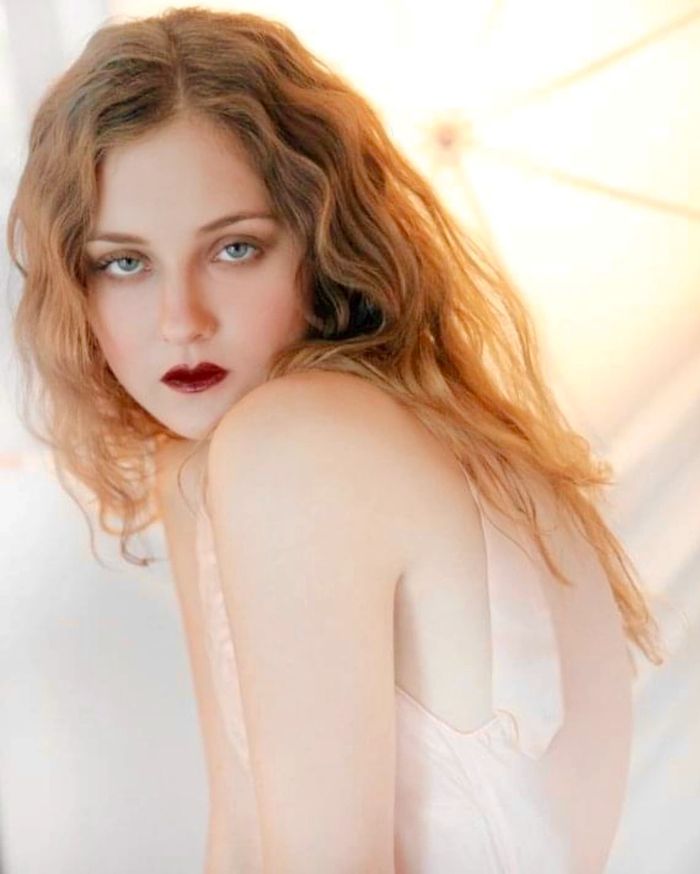 Woman with wavy ginger hair and intense blue eyes in soft lighting.
