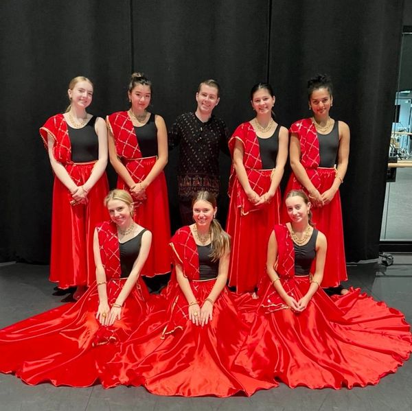 A male choreographer, surrounded by 7 young female dancers wearing vibrant red and gold tranditional