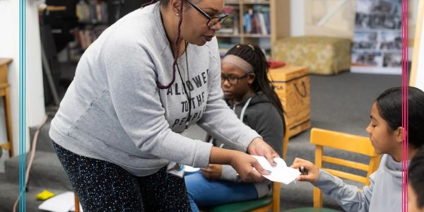 Teacher handing a paper to a student in a classroom.