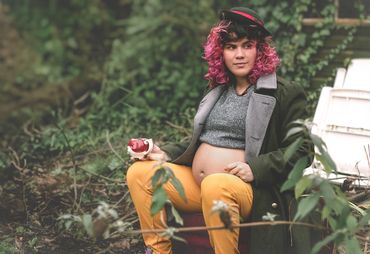 Pregnant woman with pink hair sits outdoors, holding a red teacup, wearing a coat and yellow pants.