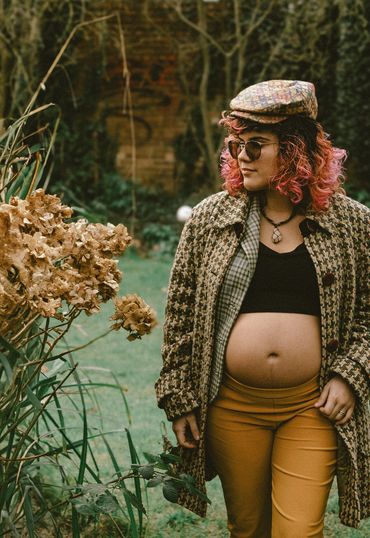 Pregnant woman in stylish autumn outfit standing outdoors near dried flowers.
