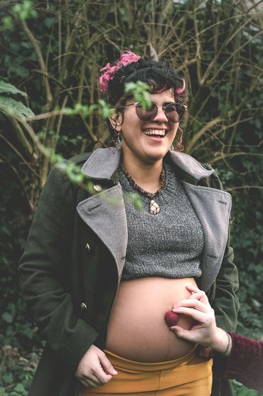 Pregnant woman smiling outdoors with a hand holding a strawberry on her belly.