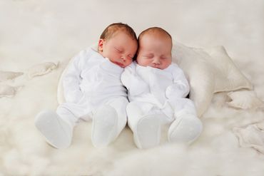 Two newborn babies sleeping peacefully side by side in white outfits.