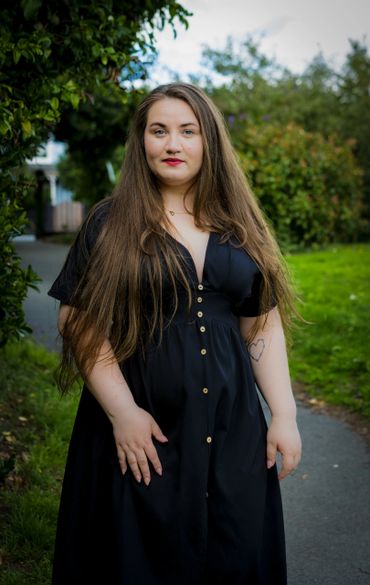Woman with long hair in a black dress standing on a pathway outdoors.
