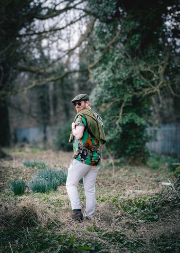 Man in colorful shirt and cap stands in a grassy, wooded area.