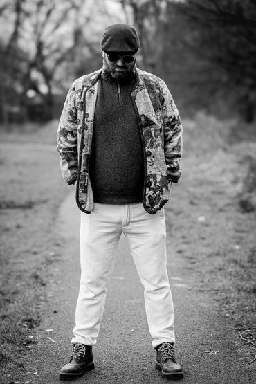 Bearded man in sunglasses and patterned jacket standing on a path outdoors.
