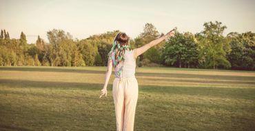 A person with feathered hair accessories stands in a sunny field pointing in two directions.