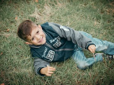 Smiling boy lying on grass holding flowers, wearing a jacket and jeans.
